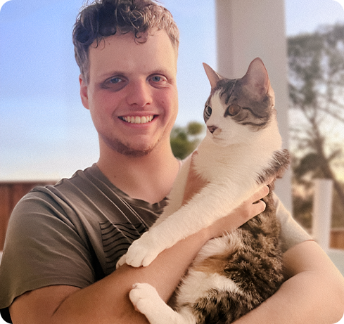 A picture of Max Lowe and his cat. He has blonde hair and blue eyes and is smiling happily while holding his cat who is staring at a different direction.